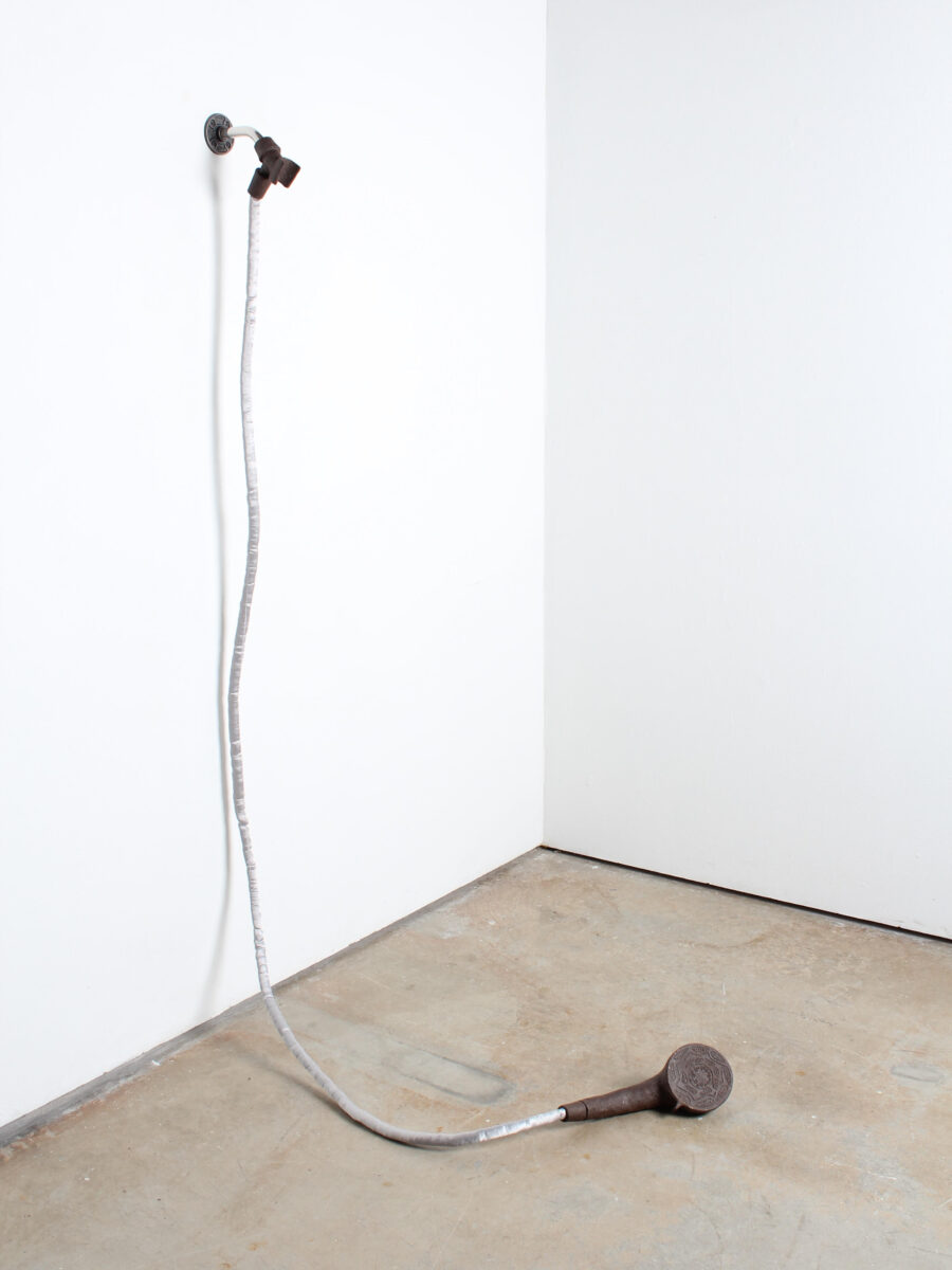 A showerhead with a fabric-wrapped hose hangs from the wall, curving down to rest on the floor in a quiet, empty corner.