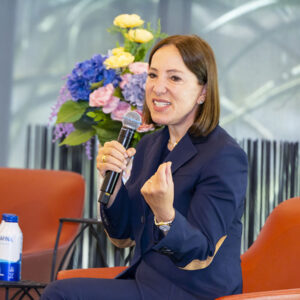 California Lieutenant Governor Eleni Kounalakis, in blue pansuit, sits in a chair and holds a microphone, gesturing with one of her hands.