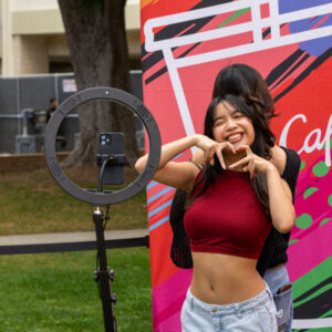 Two girls stand back to back at a 360 photo booth, one forming a heart shape with her hands.