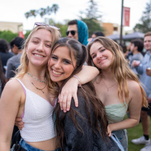 Three girls smile and pose in the middle of a crowd with their arms around each other.
