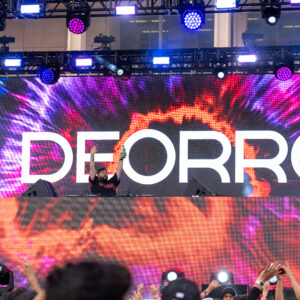 A stage with bright lights and a large screen displaying the name "Deorro" in white over a purple and orange tie-dye background.
