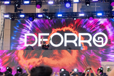 A stage with bright lights and a large screen displaying the name "Deorro" in white over a purple and orange tie-dye background.