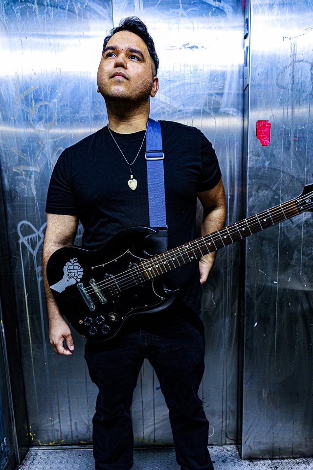 Photo of Danny Cano in an all black outfit posing with his guitar in front of a slice backdrop with white graffiti.