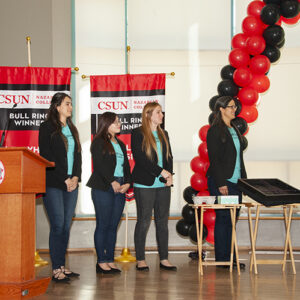 Four women stand on stage presenting their no-spill tray to the judges.