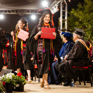 A graduate holds her red diploma cover and smiles as she crosses the commencement stage.