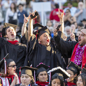 A group of four graduates in caps and gowns stand and cheer in the middle of a sea of students, with their hands in the air, at the commencement ceremony for CSUN's Mike Curb College of Arts, Media, and Communication.
