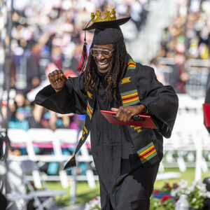 A graduate smiles and prepares to fist-bump, as he holds his red diploma cover. He is in a cap and gown, with a decorative, gold crown attached to his mortarboard.