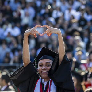 A graduate in cap and gown makes a heart symbol with two hands, holding it up in the air.