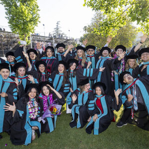 A group of about 25 doctoral graduates - in caps and gowns - celebrates by striking poses together on the Sierra Quad Lawn.