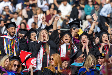A group of graduate yell and raise their fists in a commencement crowd.
