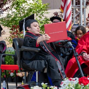 A graduate holds up his diploma as he crosses the stage in a motorized chair.