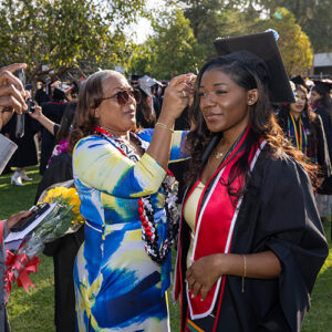 A woman helps a graduate adjust her regalia while a man holds a camera up for her to see.