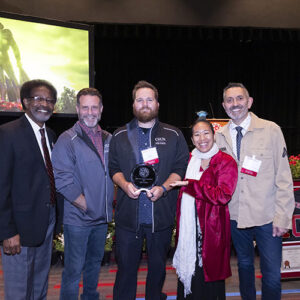 Five people pose together indoors at an awards ceremony. The person in the center holds a round glass award.