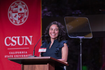 Daniela Muriel Robledo stands, smiling, at a podium. A red CSUN banner is behind her.