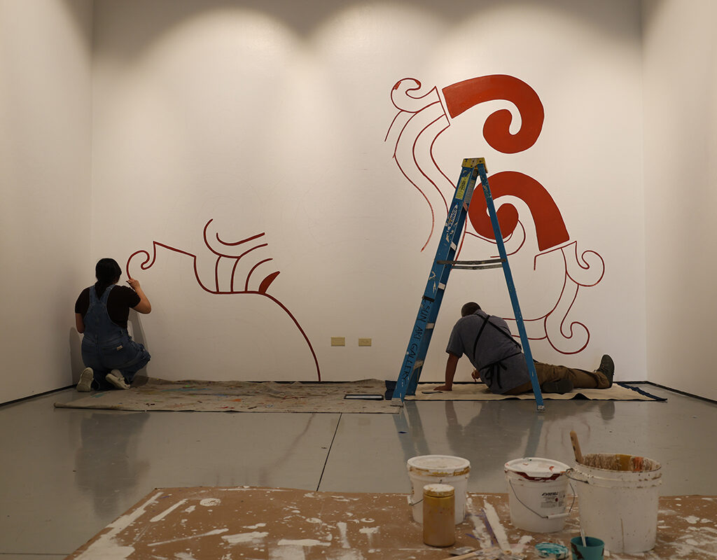 Alfonso Aceves, co-founder of Kalli Arte Collective, right, works with a student to paint a background for his residency exhibition in the CSUN Art Galleries. (Becky Savell / CSUN)
