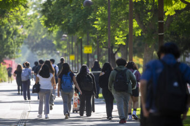 Students walk on a pathway under green, leafy trees.
