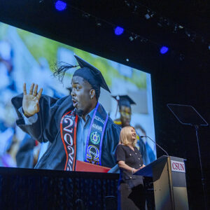 President Erika D. Beck on stage at the podium with a large screen behind her showing a celebrating graduate.