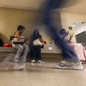 Students sit on benches in a hallway, as someone walks past in the foreground.