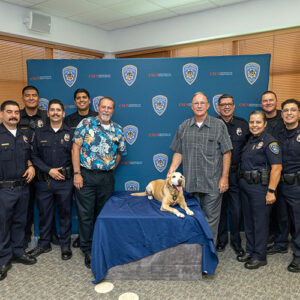 Officers dressed in black police uniforms pose against a backdrop with Randell Abney and Virgil Messmore. Golden Labrador Daisy lies on a table in the center of the group.