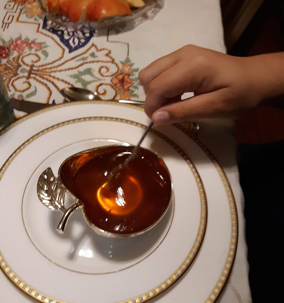 A hand dips a spoon in an apple-shaped, silver bowl of honey. A plate of apple slices is pictured in the background.