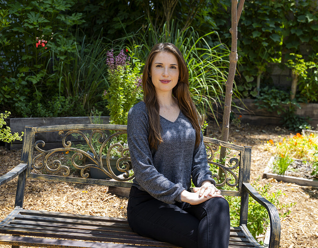 Onna Victoria Fyssicopulos seated on a bench, with plants in the background.