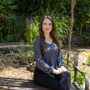 Onna Victoria Fyssicopulos seated on a bench, with plants in the background.