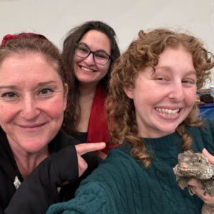 Hillel Rabbi Jess Yarkin points at a CSUN student holding a large lizard, as they pose for a selfie.