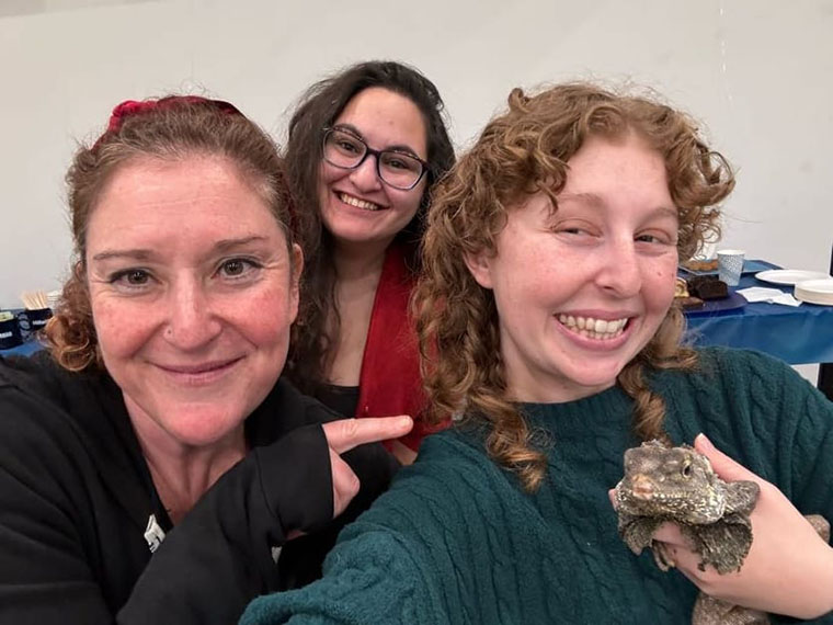 Hillel Rabbi Jess Yarkin points at a CSUN student holding a large lizard, as they pose for a selfie.