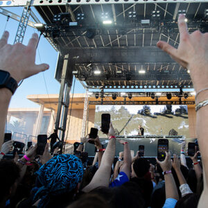In foreground, hands raised with fingers in metal horns, in background, crowd members hold up their phones.