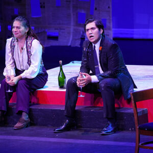 A woman and a man who are facing the audience are seated on a platform on stage.