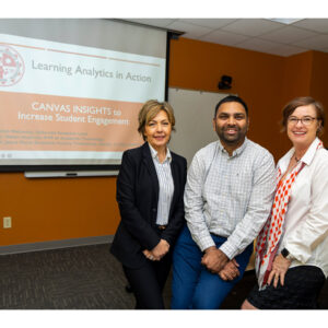Three people pose, smiling at the camera, with a video screen in the background