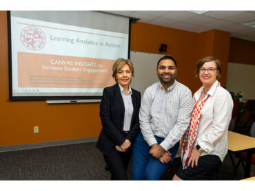 Three people pose, smiling at the camera, with a video screen in the background