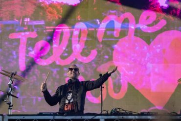 Galantis stands on stage, smiling with his arms raised, with video display in background.
