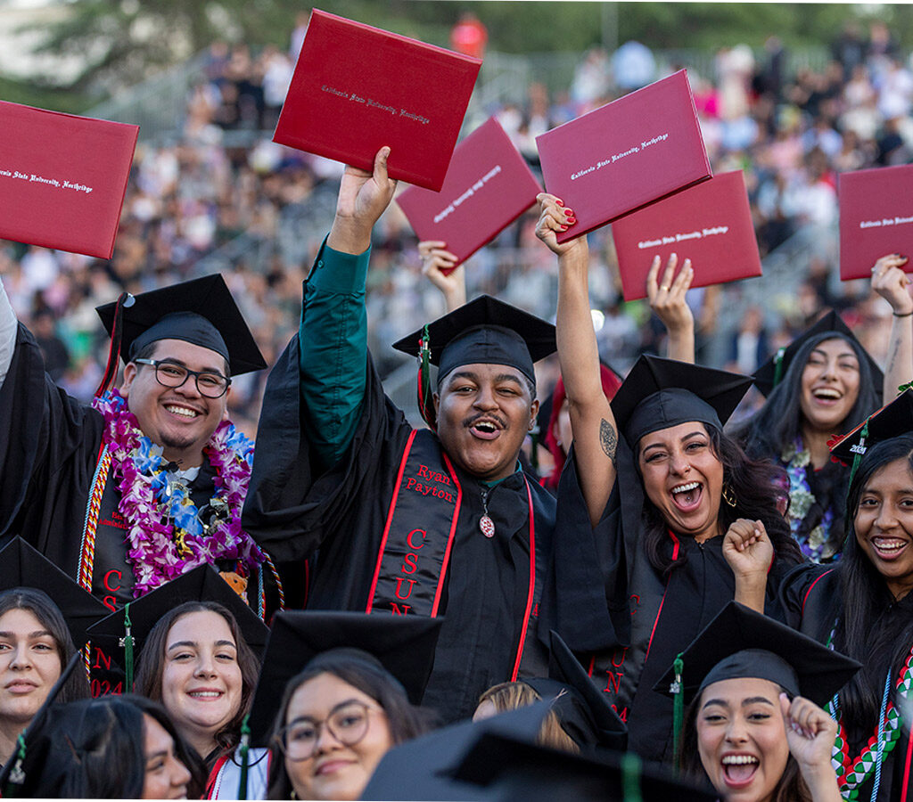Graduates in black caps and gowns hold up red diploma covers.