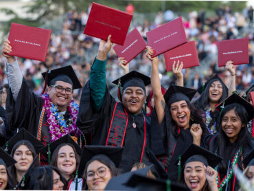 Graduates in black caps and gowns hold up red diploma covers.