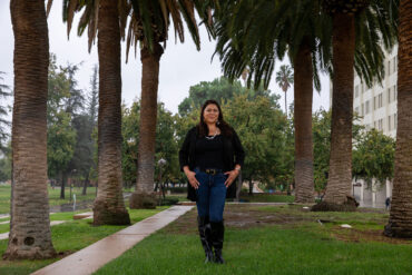 Jenee Ornelas' stands in the rain between rows of palm trees in front of the University Library.