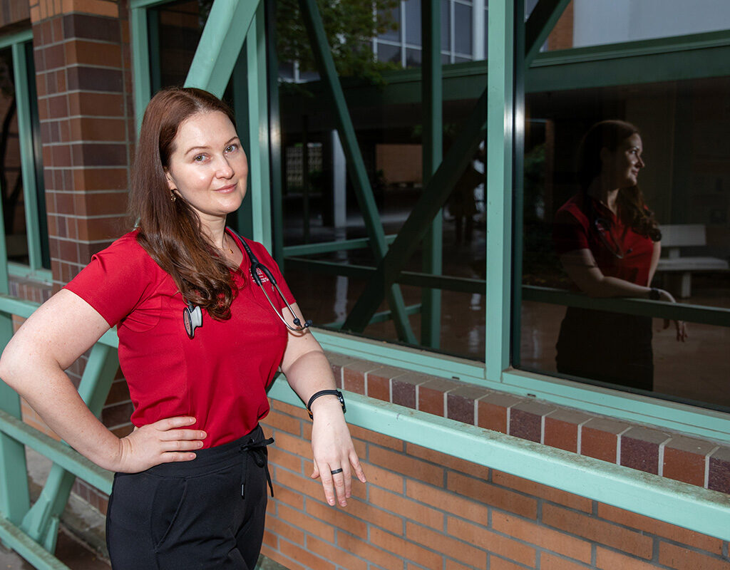 Nursing student Luba Buklemisheva, wearing a red shirt, stands in front of a window. 