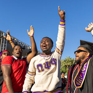 Graduates, wearing fraternity letters, raise their arms in the air.