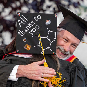 A student with mortarboard that says "All Thanks to You" hugs a smiling professor.