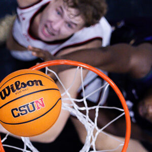 A basketball with "Wilson CSUN" hovers over the basket with two players below.