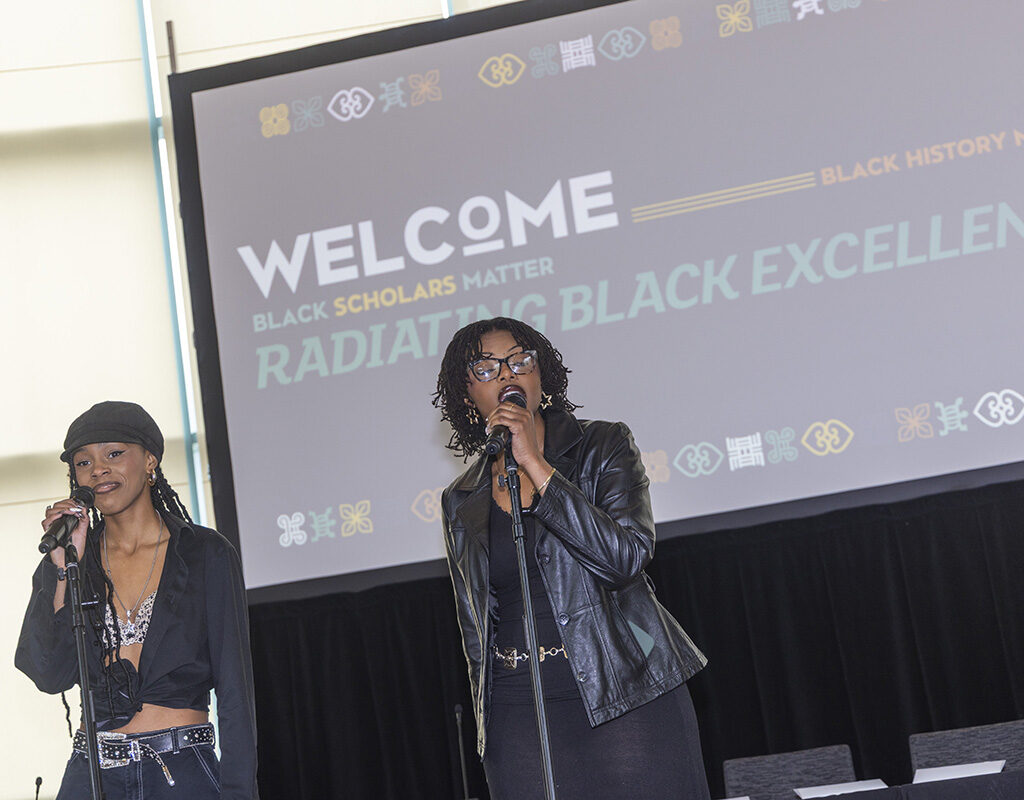 Two women stand on stage with microphones, video screen in the background.