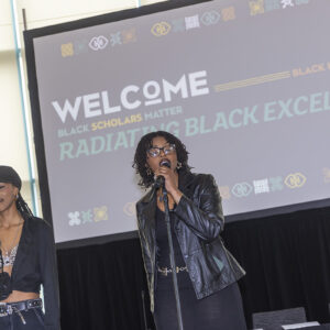 Two women stand on stage with microphones, video screen in the background.