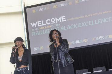 Two women stand on stage with microphones, video screen in the background.
