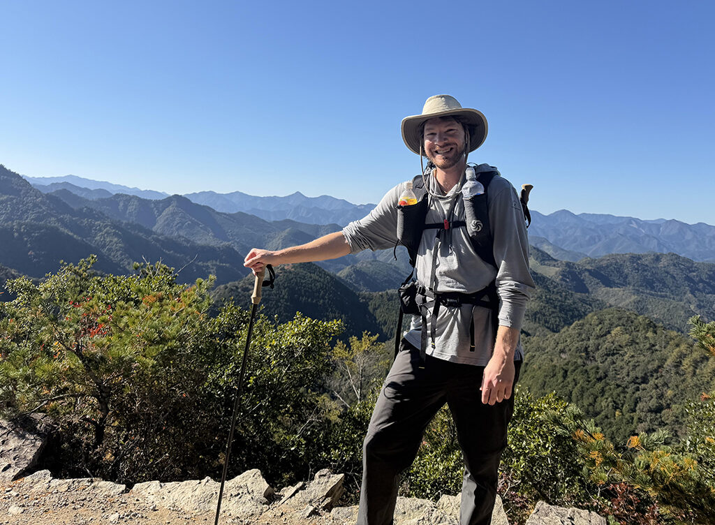 A man at a high point on a wilderness hike with trees and mountains in the distance.