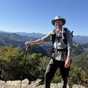 A man at a high point on a wilderness hike with trees and mountains in the distance.