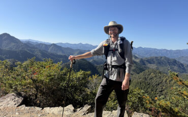 A man at a high point on a wilderness hike with trees and mountains in the distance.