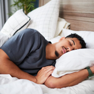 A young man is resting soundly on a cozy bed, embracing a pillow while surrounded by soft white linens, captured in a serene and calm moment within a home setting.