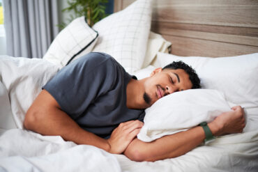 A young man is resting soundly on a cozy bed, embracing a pillow while surrounded by soft white linens, captured in a serene and calm moment within a home setting.