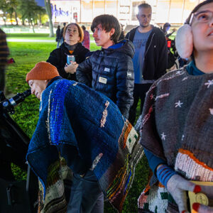 People gathered on the University Library Lawn at night look through a telescope; a person peers into it, while others look up at the sky.