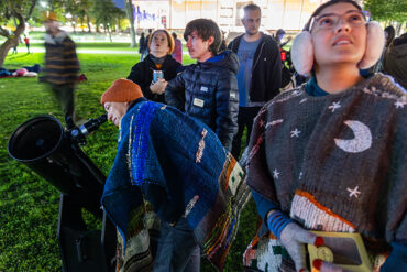 People gathered on the University Library Lawn at night look through a telescope; a person peers into it, while others look up at the sky.